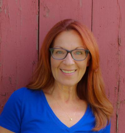 Woman with red hair and glasses, wearing a blue shirt, standing against a red wall